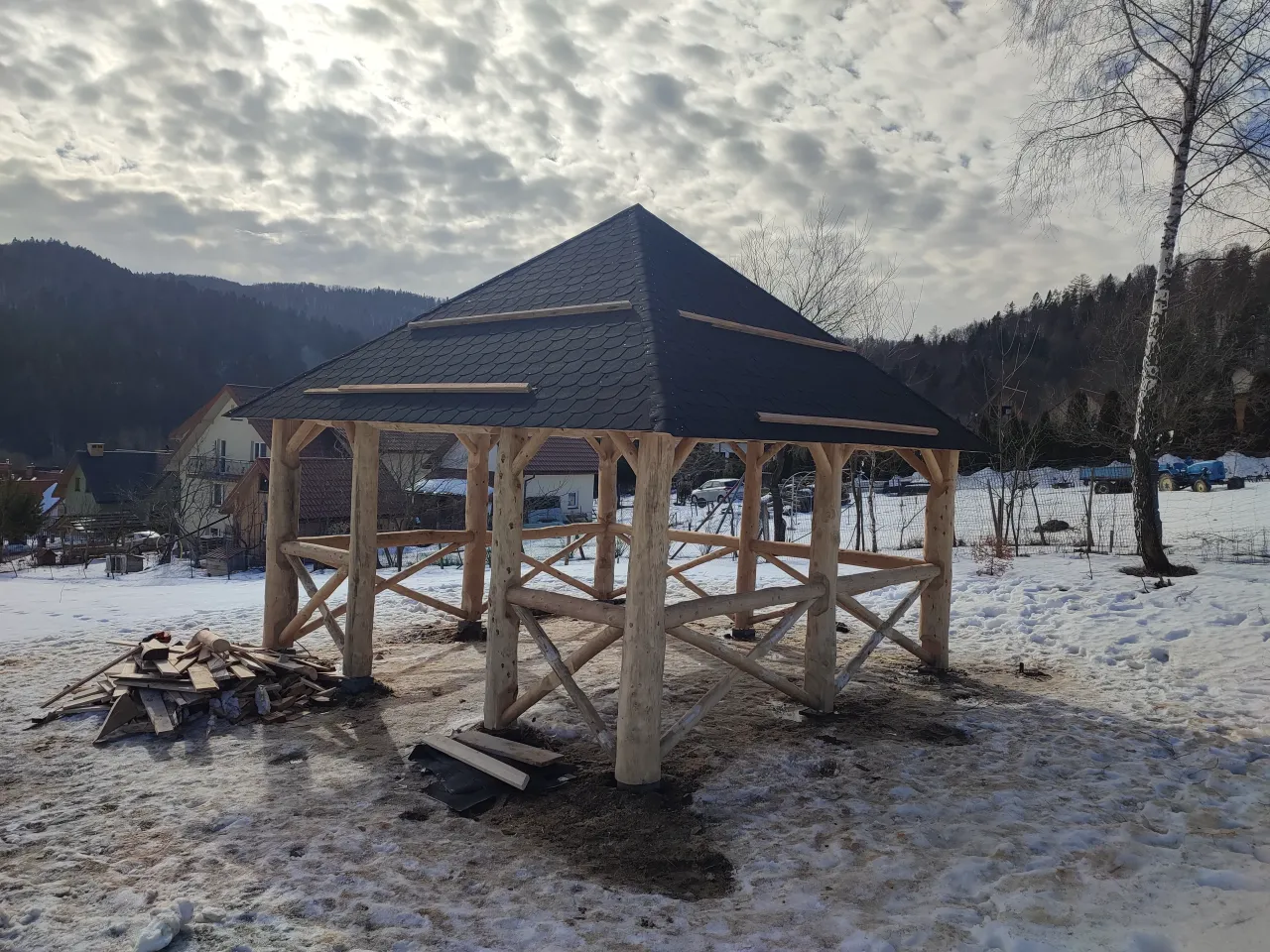 Drewniana altana w trakcie budowy, z ciemnym dachem z gontu. Wok&oacute;ł śnieg, stos desek. W tle g&oacute;ry i domy.
