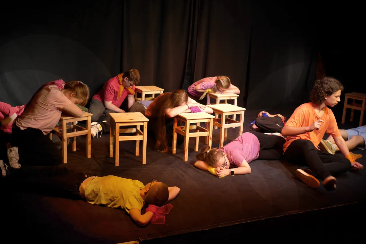 Dzieci na scenie, w ramach przedstawienia teatralnego, leżą lub pochylają się nad drewnianymi stołkami, tworząc scenę pełną emocji.