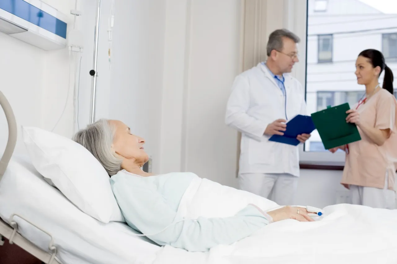 Starsza pacjentka w ł&oacute;żku szpitalnym, lekarz i pielęgniarka omawiający jej stan. Wszyscy starają się minimalizować czynniki jatrogenne.