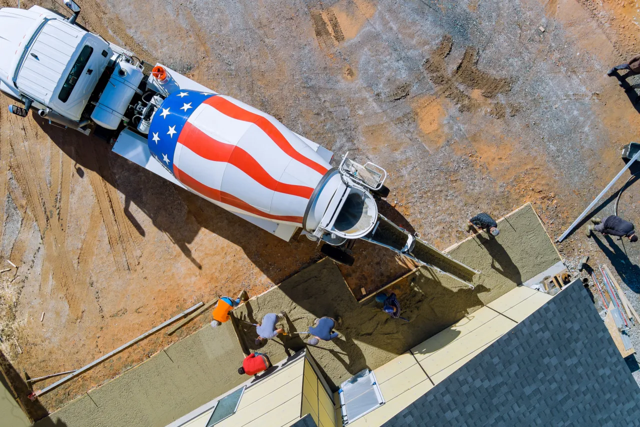 Betonowanie fundamentów domu. Z betoniarki z amerykańską flagą wylewany jest beton. Pracownicy rozprowadzają go łopatami.