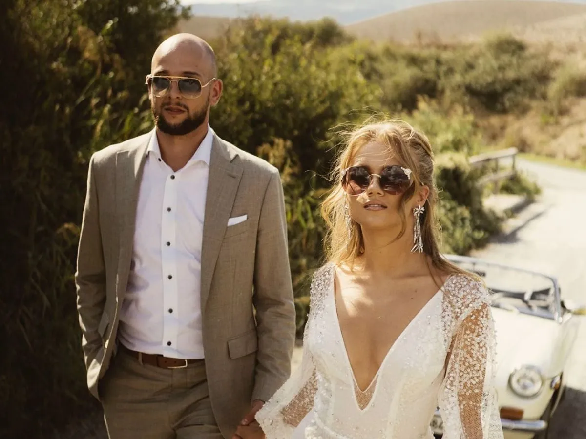 Agnieszka Kaczorowska i jej mąż idą trzymając się za ręce. Ona w sukni ślubnej, on w garniturze. W tle zabytkowy samochód.