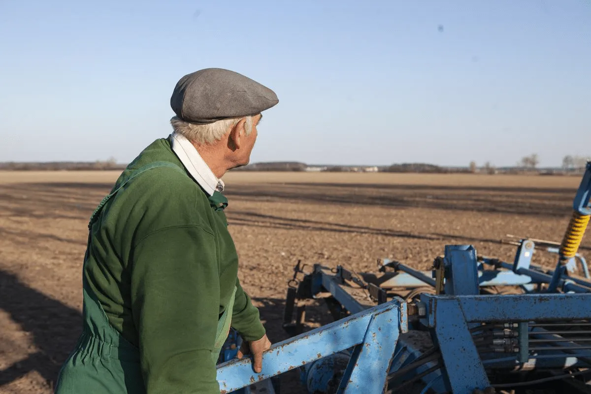 Rolnik w zielonym ogrodniczkach i czapce na tle pola, zastanawia się nad wiekiem emerytalnym rolników.