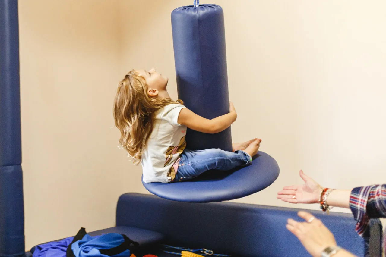 Dziecko z zaburzeniami sensorycznymi huśta się na niebieskim przyrządzie. Terapia sensoryczna pomaga w rozwoju.