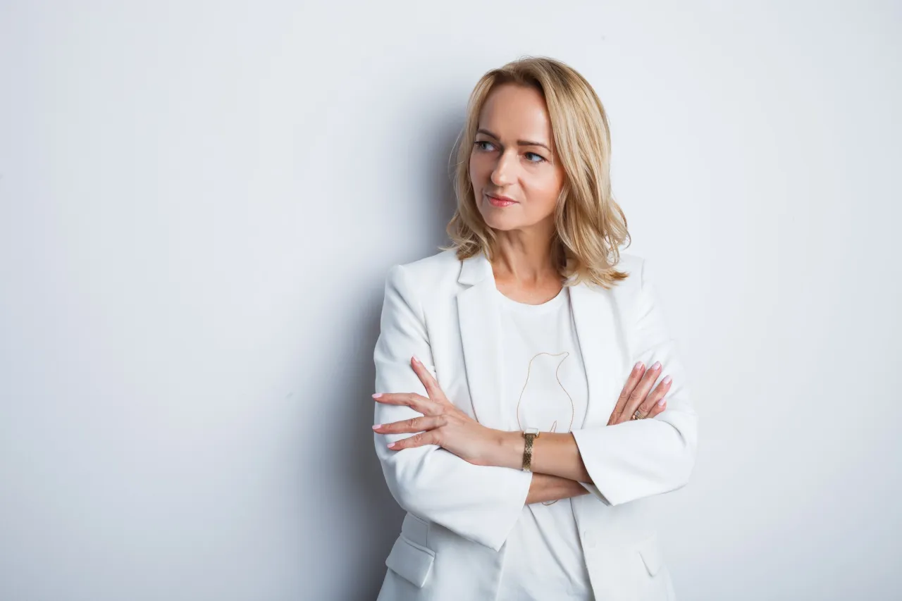 Kobieta w białej marynarce, z rękami skrzyżowanymi na piersi, patrzy w bok. Idealny przykład, jak zrobić zdjęcie do CV telefonem.