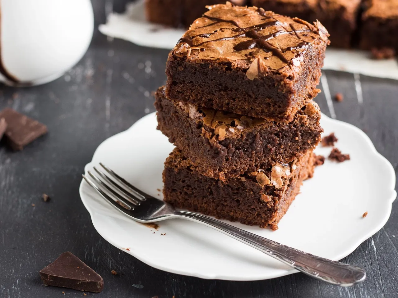 Pyszne, wilgotne brownie ułożone na talerzyku, obok widelca i kawałk&oacute;w czekolady. Idealny przepis na zdrowe ciasto.
