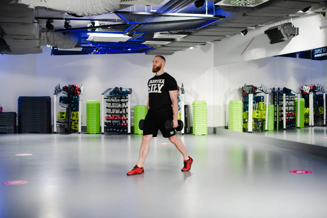 Mężczyzna wykonuje przysiad z hantlami w siłowni. W tle stojaki ze sprzętem i lustra.