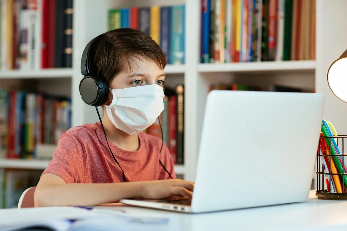 Chłopiec w masce i słuchawkach uczy się zdalnie przy laptopie. Na półkach za nim widać książki.