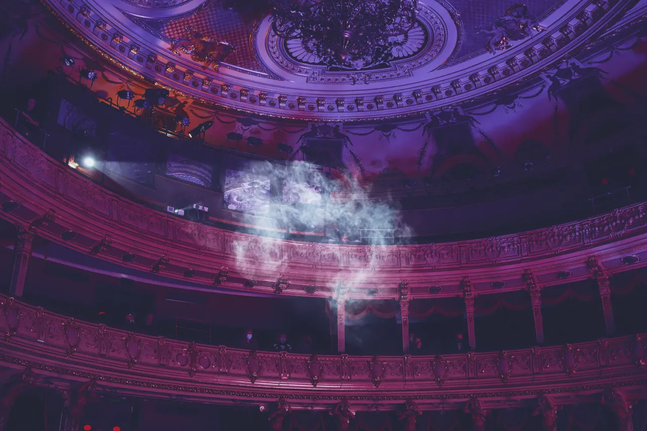 W teatrze, pośród dymu i purpurowego światła, widzimy bogato zdobione loże i widownię. To miejsce, gdzie rodzą się emocje i opowieści.