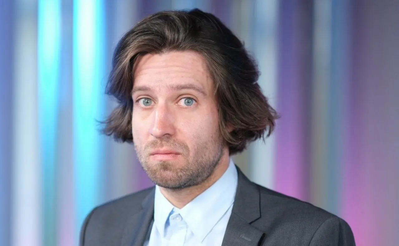 Michał Kempa stand up, w garniturze i jasnej koszuli, z burzą brązowych włos&oacute;w, patrzy w obiektyw na tle kolorowych pas&oacute;w.