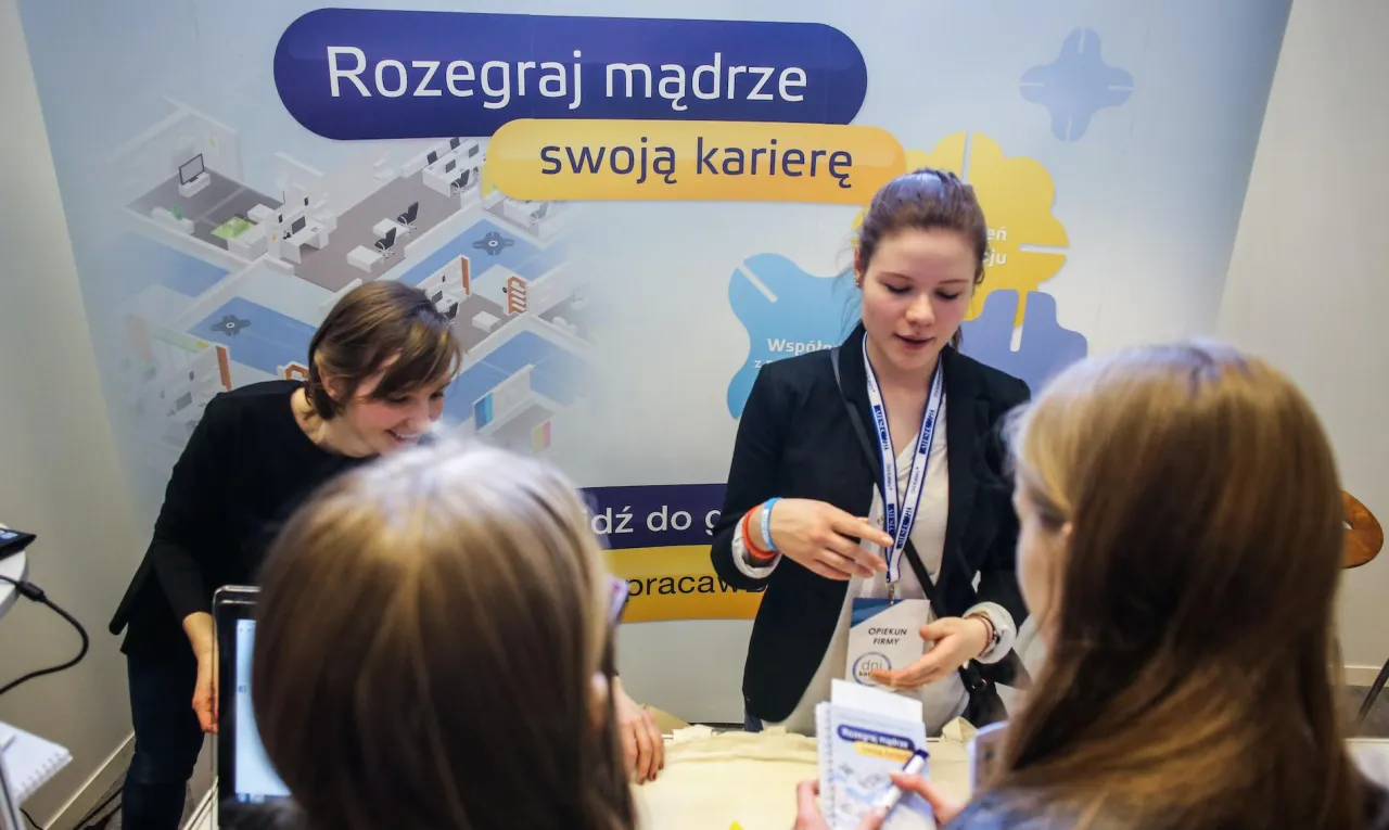 Młode kobiety rozmawiają przy stoisku informacyjnym. Dowiadują się, jak przygotować się do pracy w branży marketingowej, analizując materiały.