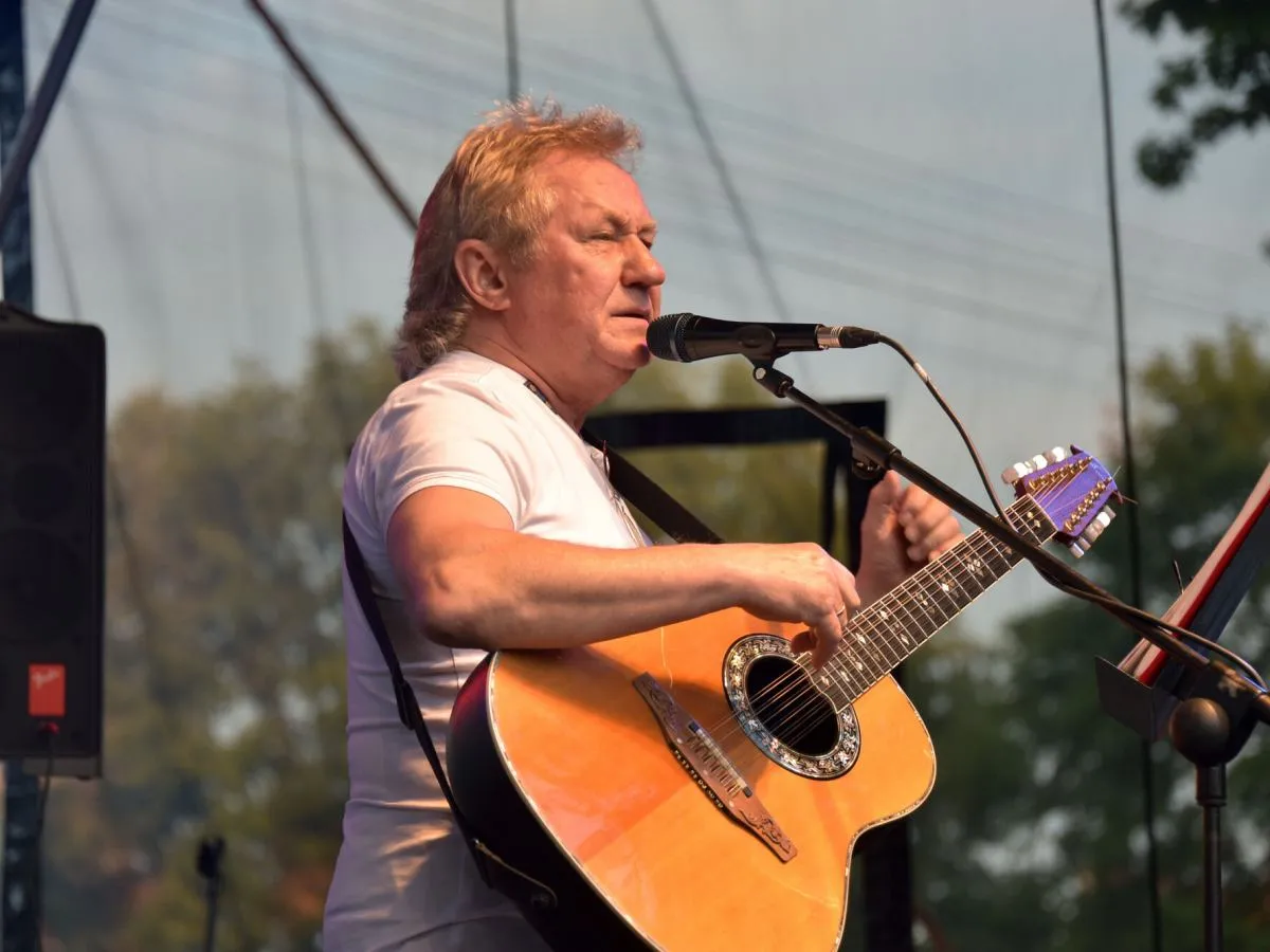 Artysta z blond włosami gra na gitarze, śpiewając na scenie. Kto pierwszy śpiewał kolorowe jarmarki?