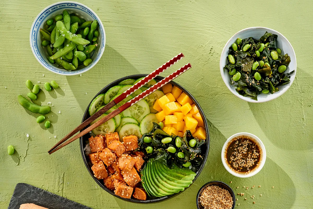 Kolorowa miska poke z łososiem, awokado, mango, og&oacute;rkiem i edamame. Obok miseczka z sałatką wakame, idealna do tego wakame przepis.