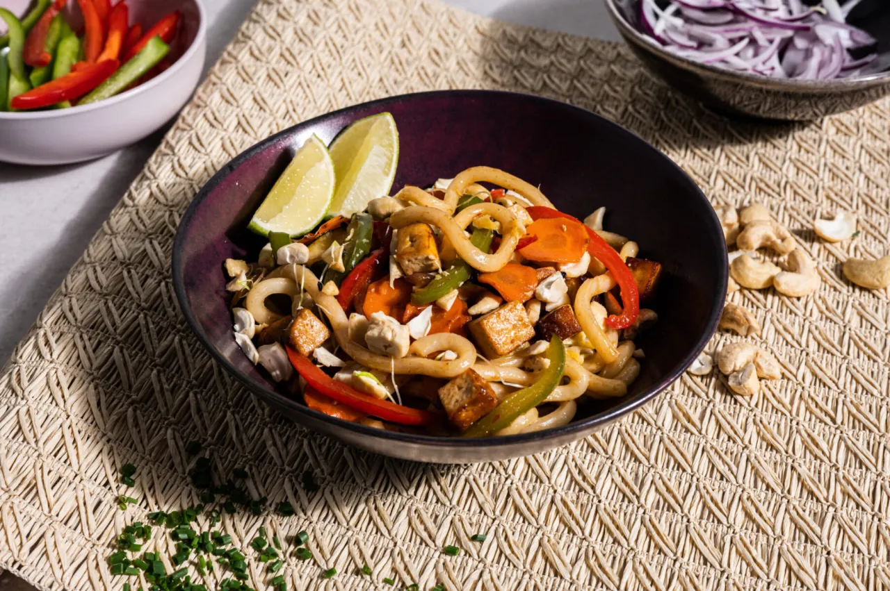 Pyszny udon przepis z tofu, warzywami i orzechami nerkowca. Danie podane z limonką i posypane szczypiorkiem.