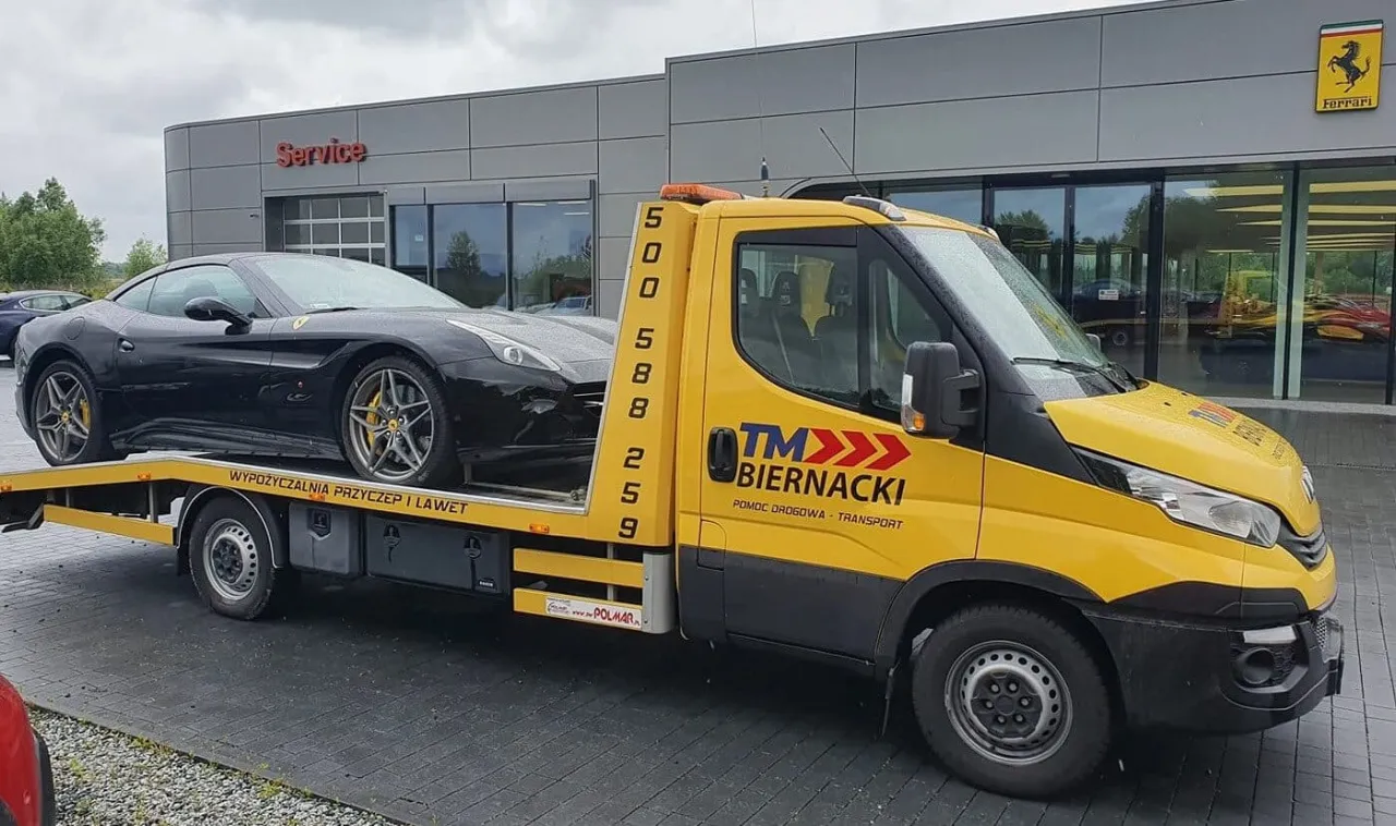 Ż&oacute;łta laweta z logo TM Biernacki zapewnia pomoc drogową dla czarnego Ferrari.