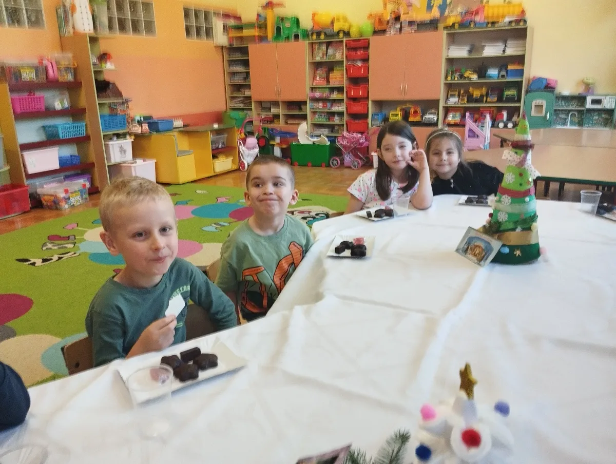 Dzieci w przedszkolu, gdzie zerówka jest obowiązkowa, jedzą słodycze przy stole ozdobionym choinką.