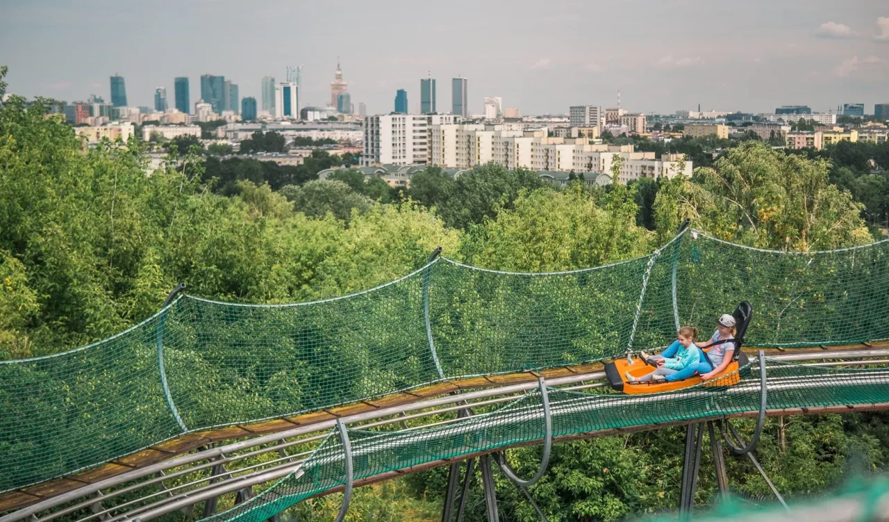 Dwie dziewczynki zjeżdżają na sankach w d&oacute;ł po torze z widokiem na panoramę Warszawy. Świetny pomysł, co zwiedzić w Warszawie z dziećmi!