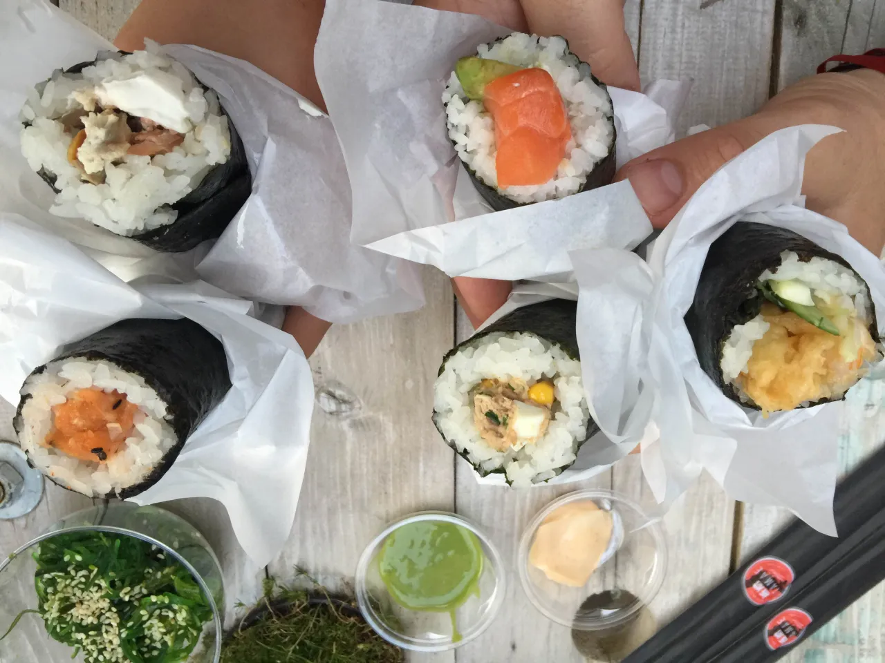 Ręcznie robione sushi w papierowych opakowaniach, z łososiem, awokado, tuńczykiem i krewetkami. Obok sosy i sałatka wakame.