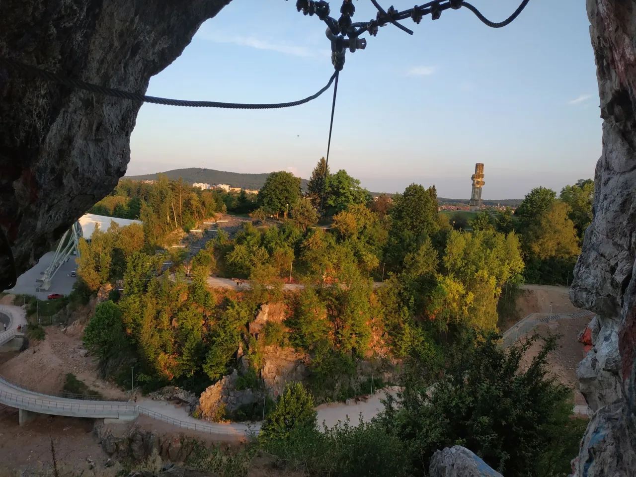 Widok z jaskini na zalesiony teren z mostami i wieżą. Rezerwat przyrody Kadzielnia w słońcu.