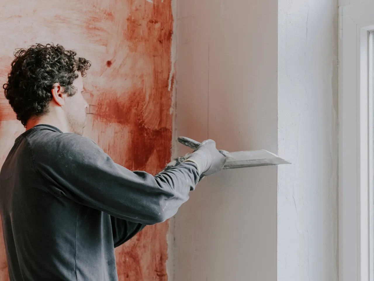 Mężczyzna w rękawiczkach nakłada masę na ścianę, przygotowując gruntowanie ścian i sufitów przed tynkowaniem.