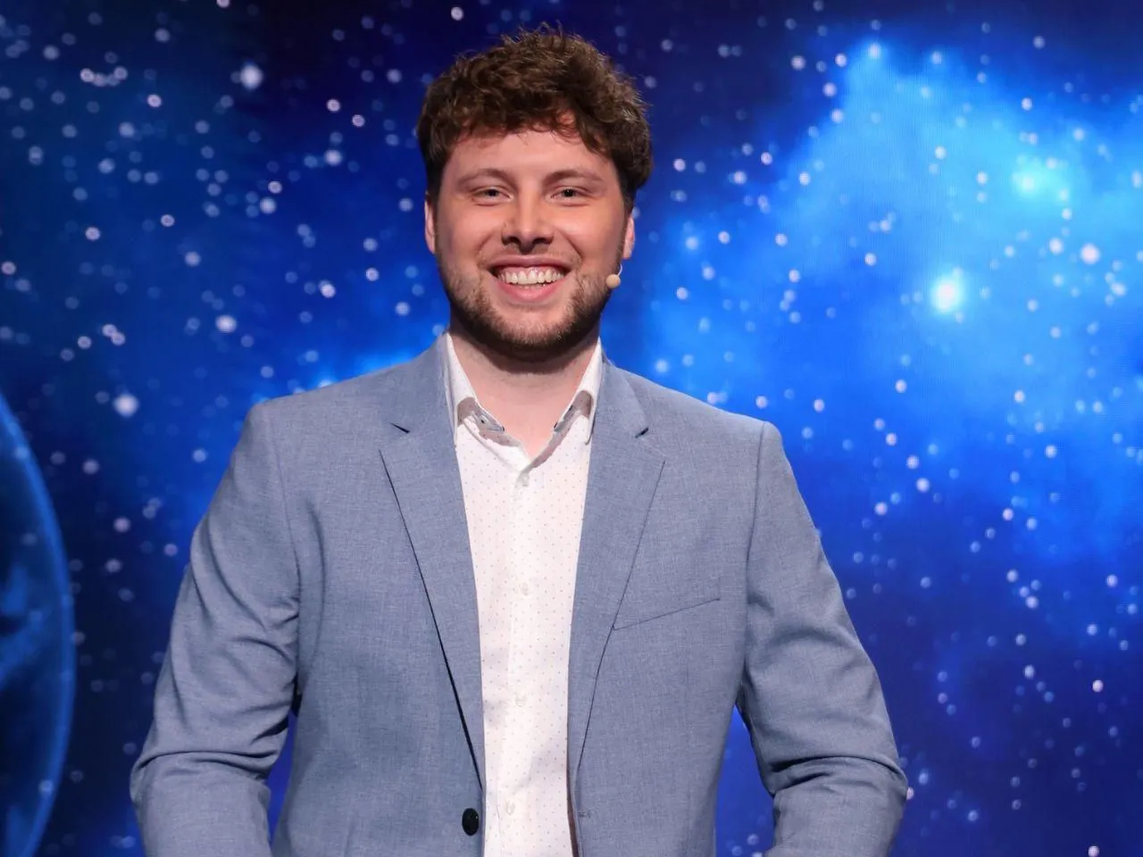 Uśmiechnięty mężczyzna w jasnej marynarce, jeden z dziesięciu jak się zgłosić, na tle kosmicznej scenografii.