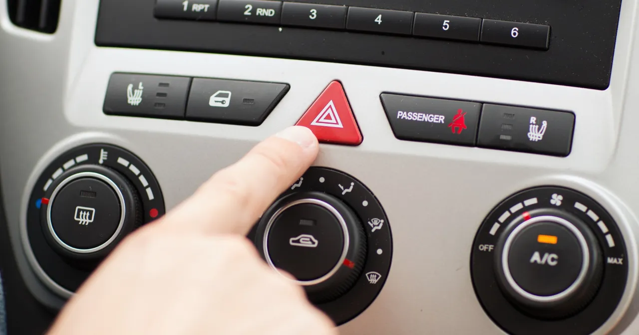 Palec naciska czerwony przycisk z trójkątem, aktywując światła awaryjne. Widać też pokrętła klimatyzacji.
