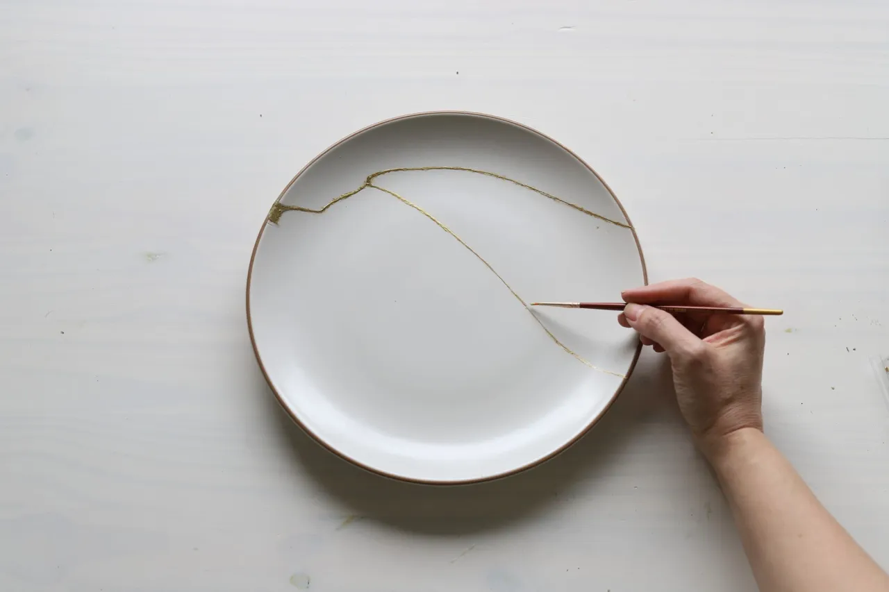Ręka pędzlem maluje złote linie na pęknięciach talerza, pokazując, jak naprawić wyszczerbiony kubek.