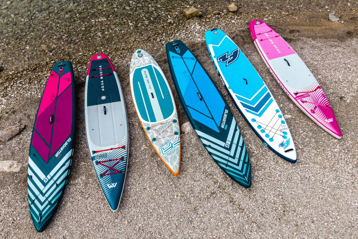 Sześć dmuchanych desek do pływania w r&oacute;żnych kolorach i wzorach leży na kamienistej plaży, gotowych na wodne przygody.