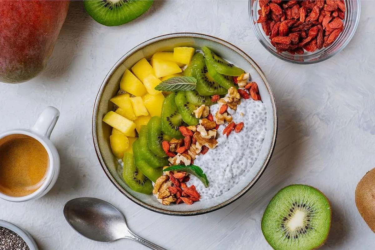 Pudding chia z mango, kiwi, orzechami i jagodami goji – idealny posiłek przed treningiem. Obok espresso i świeże owoce.