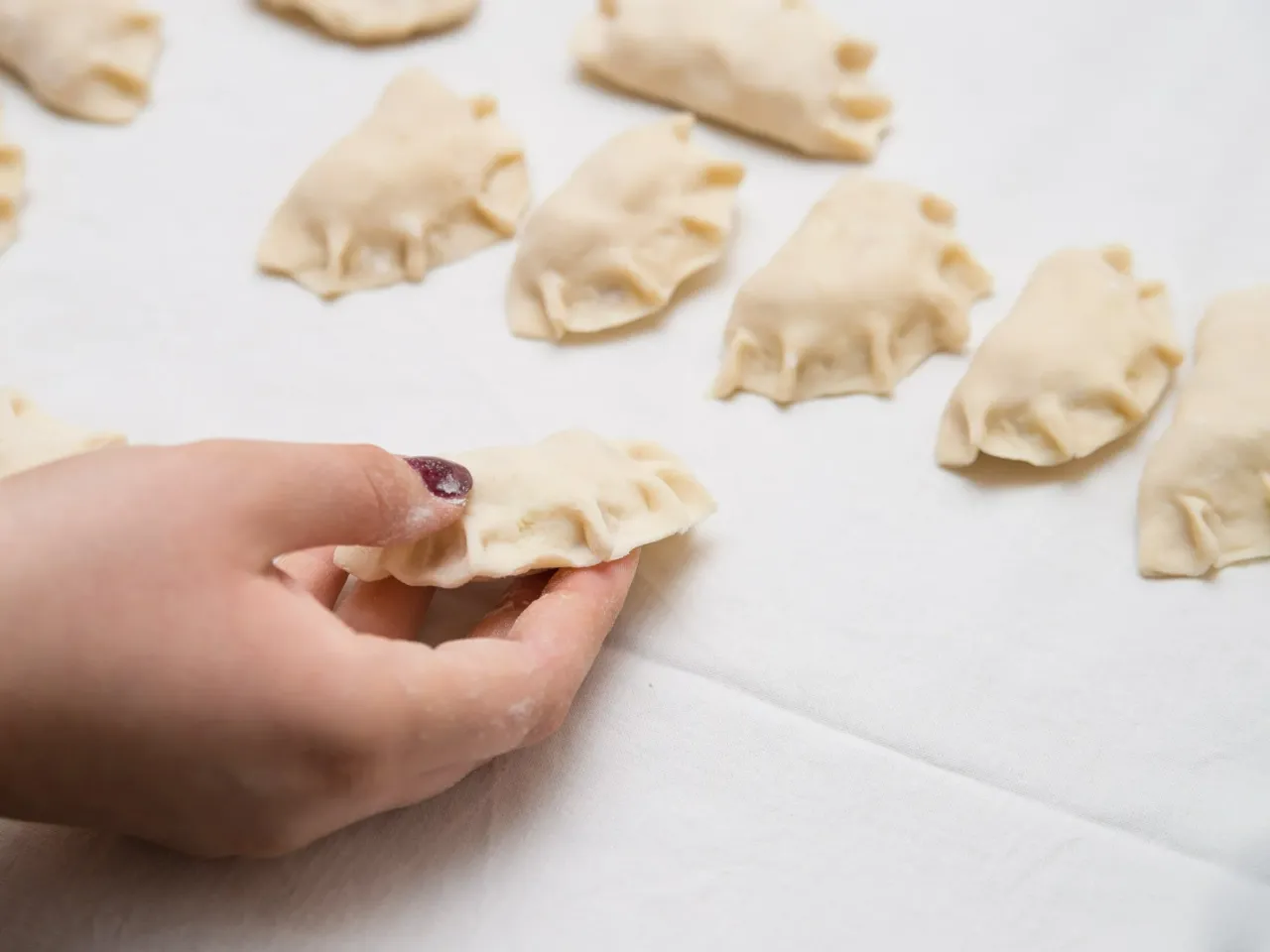 Dłoń trzyma surowe pierogi, pokazując, jak zrobić pierogi. W tle widać więcej gotowych do gotowania pierog&oacute;w.