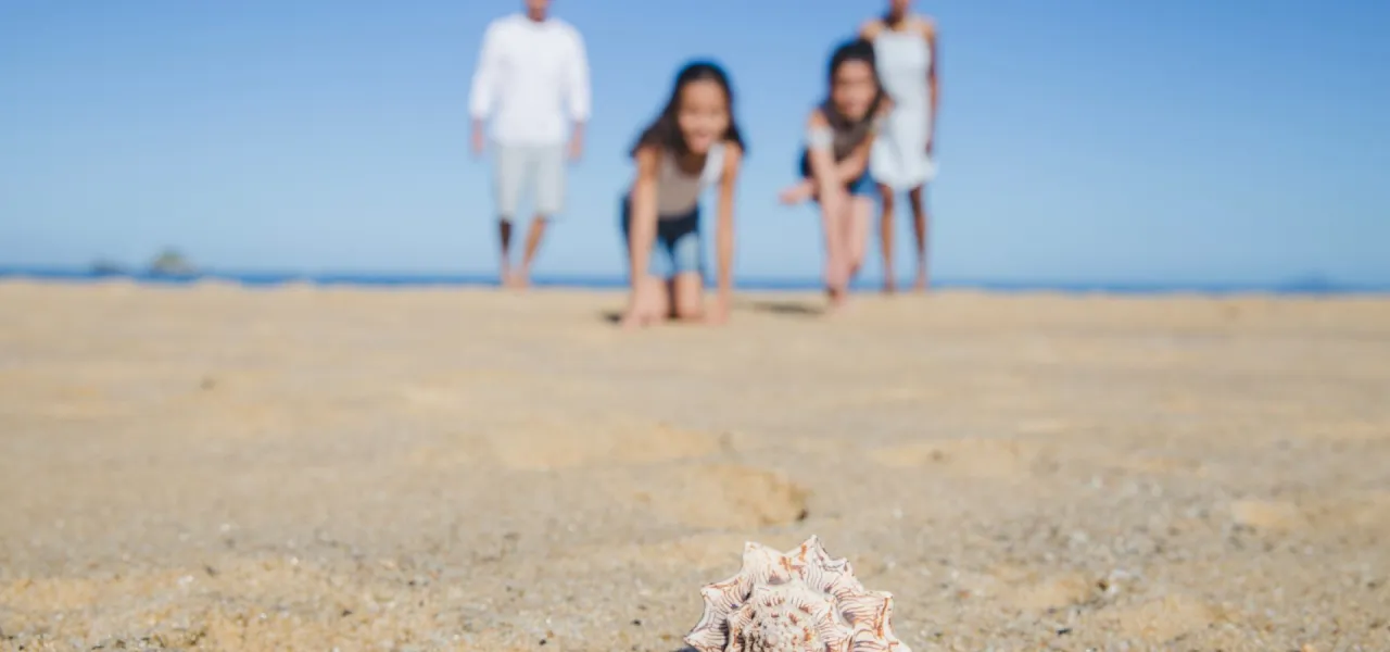 Rodzinne wakacje z dzieckiem nad morzem. Dzieci na plaży pochylone nad muszlą, rodzice w tle.