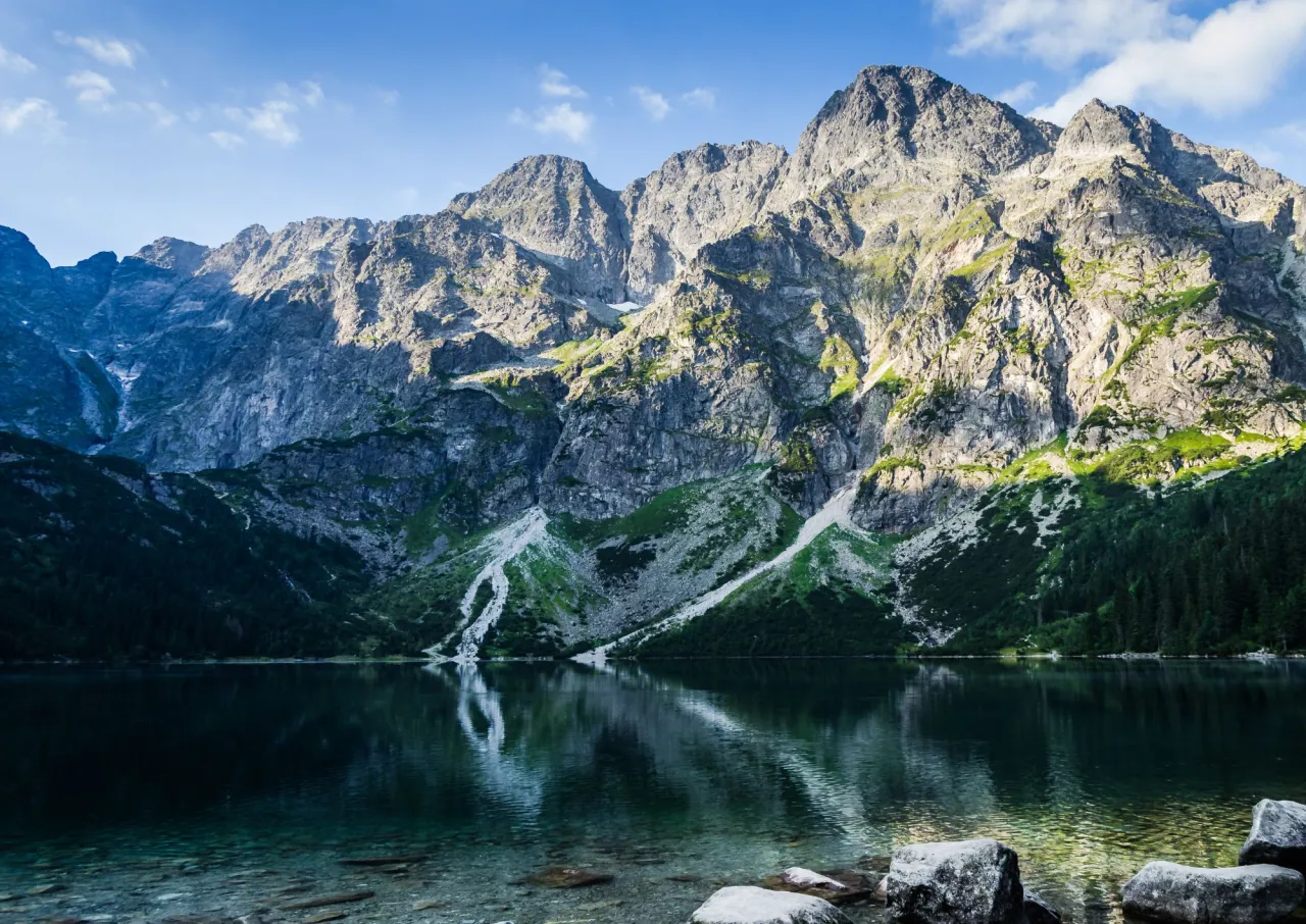Majestatyczne góry odbijają się w spokojnej tafli jeziora Morskie Oko. Trasa widoczna na zboczu zaprasza do wędrówki.