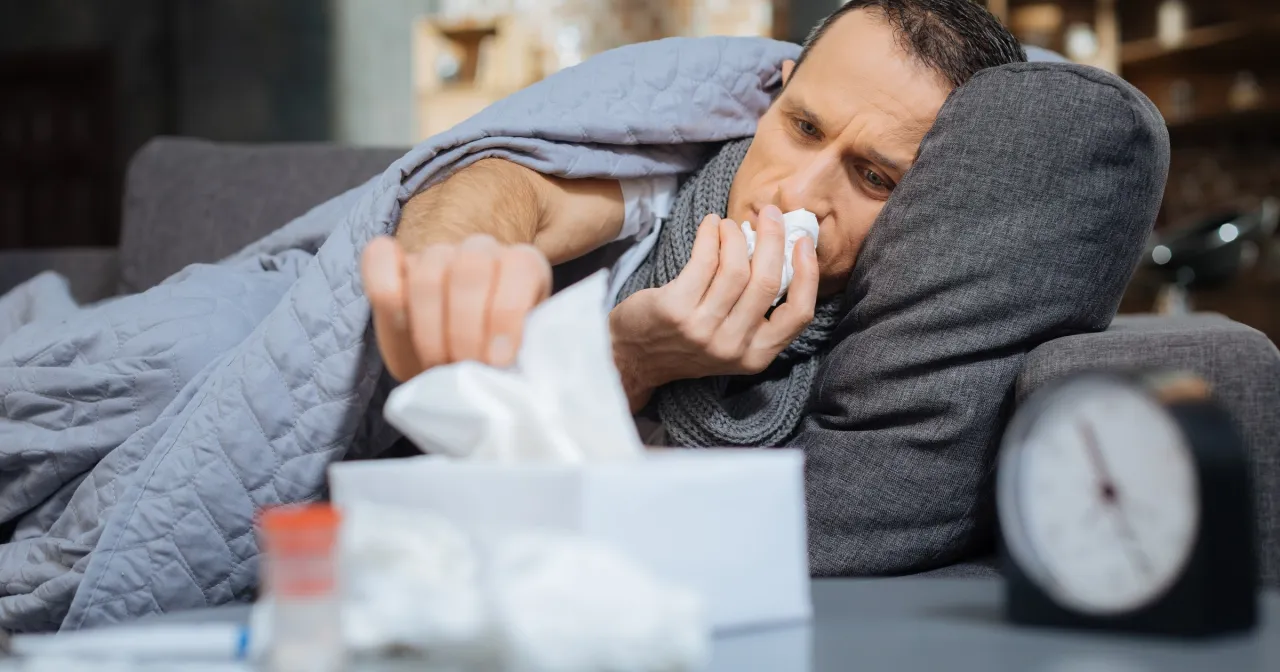 Chory mężczyzna z chusteczką przy nosie, otulony kocem. Objawy zapalenia płuc mogą być podobne do przeziębienia.