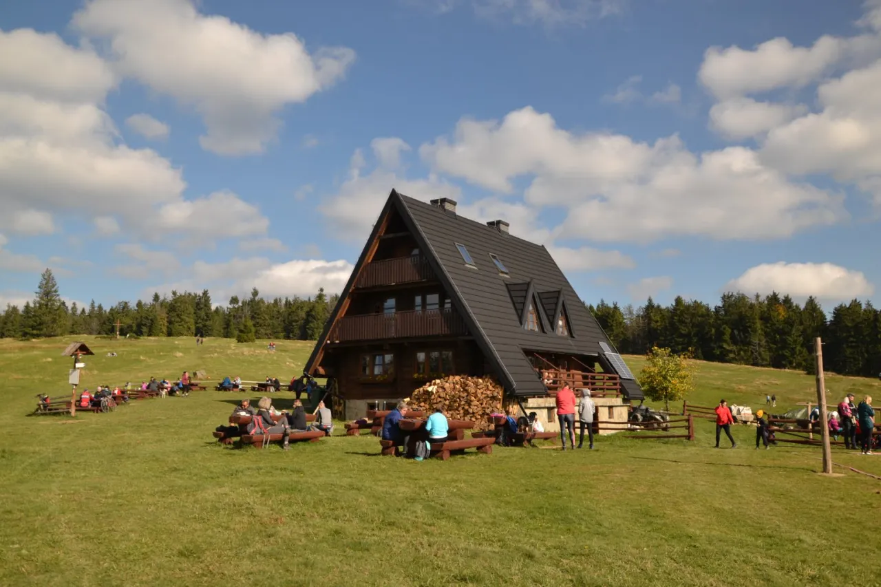 Ludzie odpoczywają przy bac&oacute;wce Krawc&oacute;w Wierch. Drewniany budynek z ciemnym dachem i zielony trawnik tworzą malowniczy krajobraz.