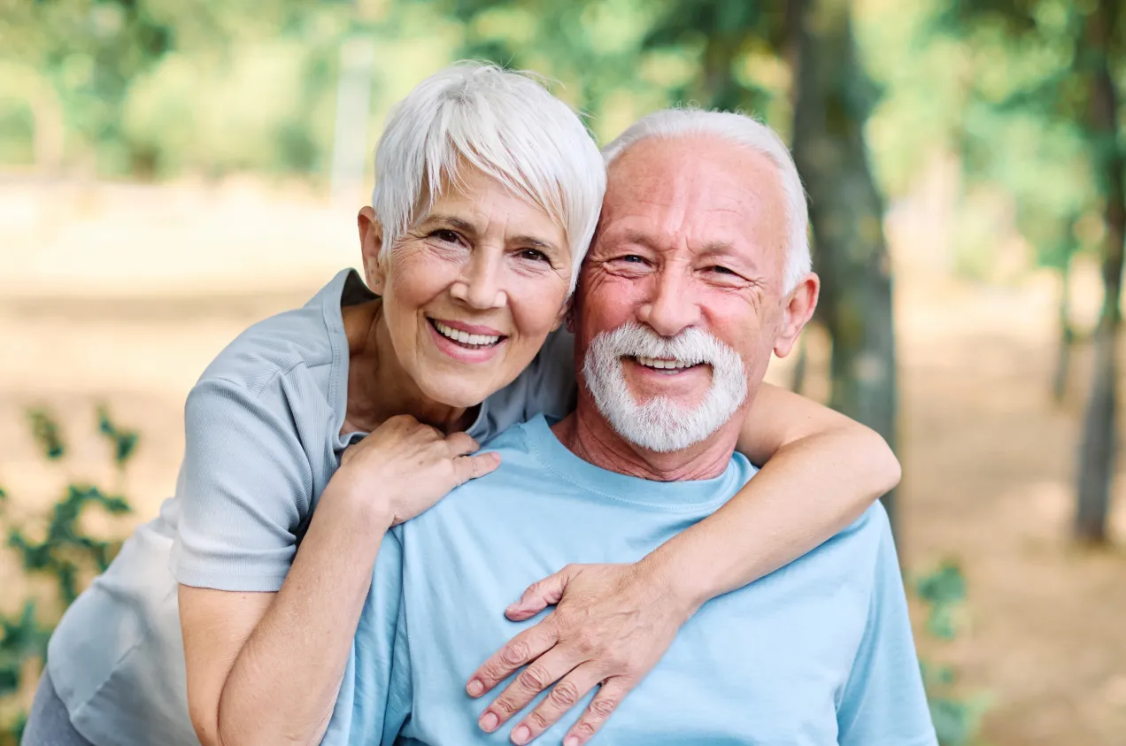 Uśmiechnięta para seniorów, ona obejmuje go ramionami. To jest właśnie obraz tego, kto to jest senior obecnie – pełen życia i radości.