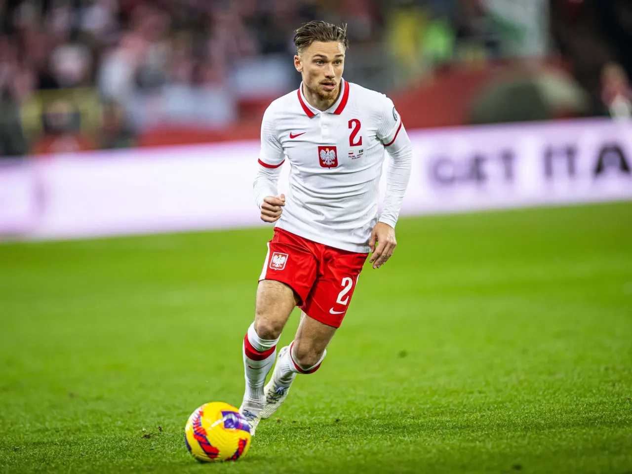 Piłkarz w białej koszulce z numerem 2 i czerwonych spodenkach, z orłem na piersi, biegnie z piłką. Matty Cash wzrostu nie przeszkadza mu w dynamicznej grze.