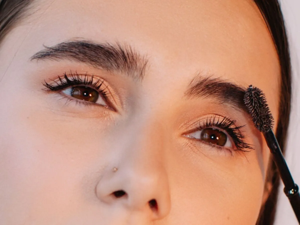 Zbliżenie na oczy z ciemną oprawą, podkreślone tuszem. Szczoteczka do brwi delikatnie modeluje łuk.