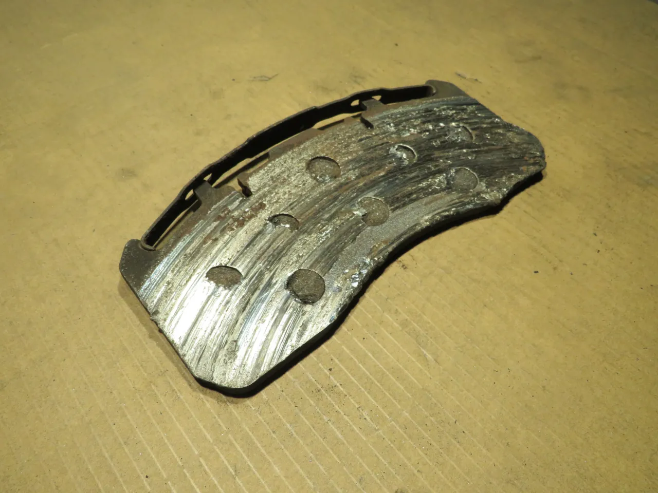 Zużyty klocek hamulcowy, z widocznymi śladami tarcia i uszkodzeń. Jego stan świadczy o niskiej żywotności klock&oacute;w hamulcowych.