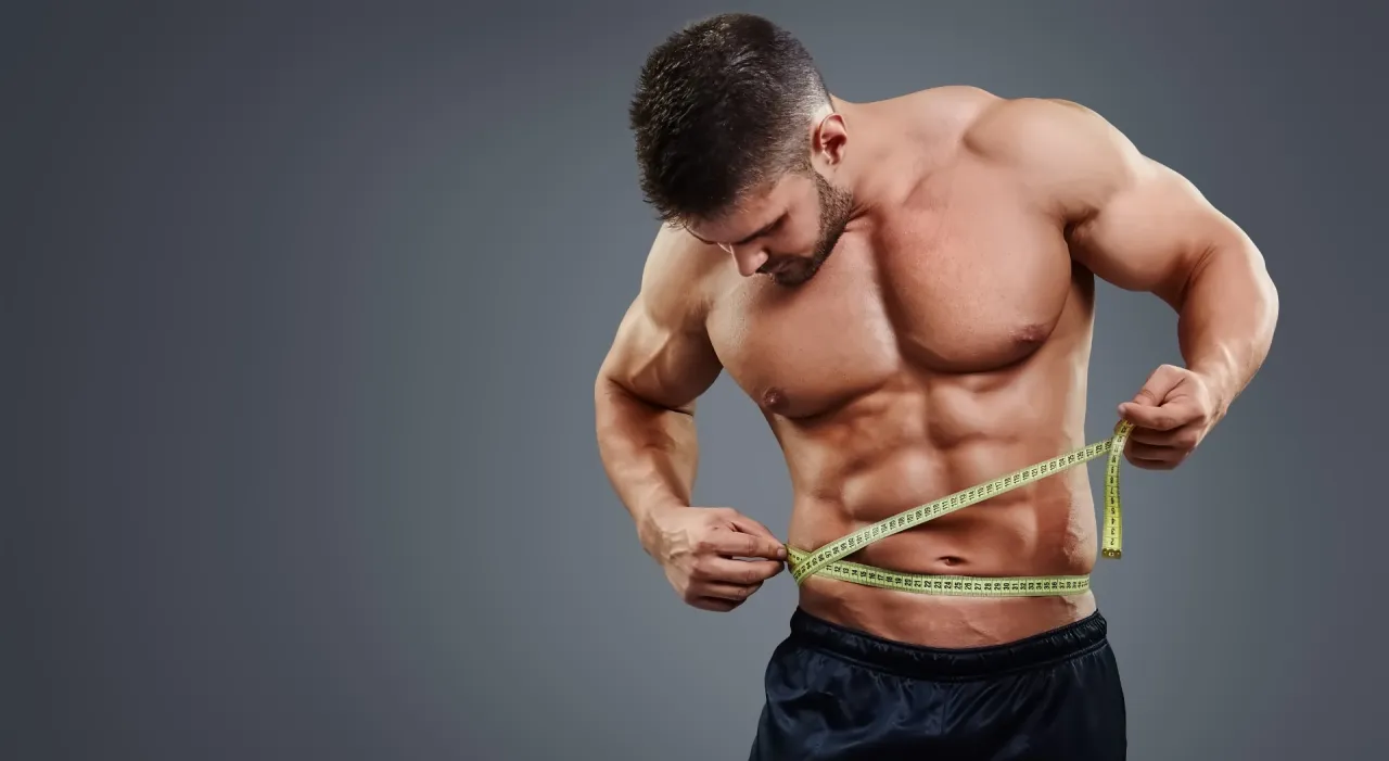 Mięśniowy mężczyzna mierzy talię centymetrem, dowodząc, że trening siłowy spala tłuszcz.