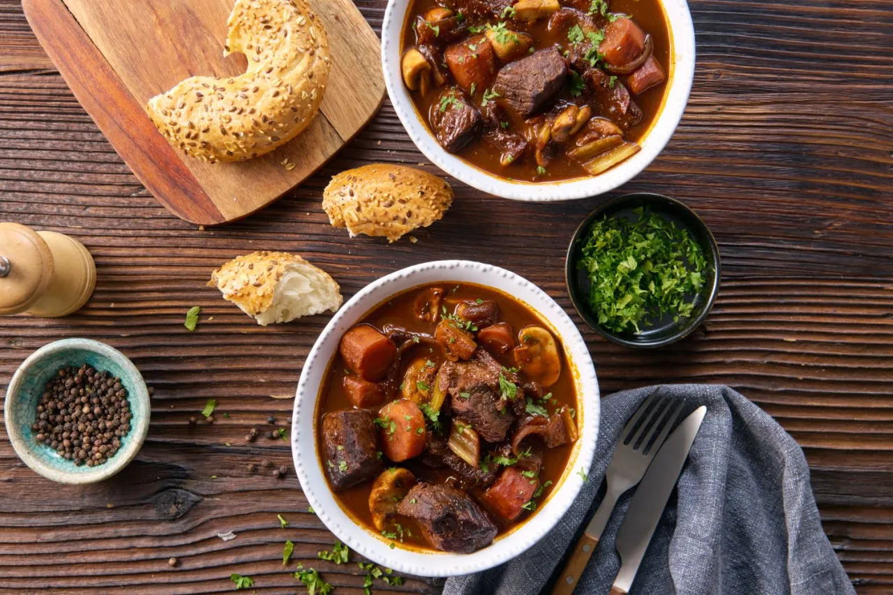 Dwa talerze aromatycznej wołowiny po burgundzku z klopsami, marchewką i grzybami, podane z pieczywem i posiekaną natką pietruszki.