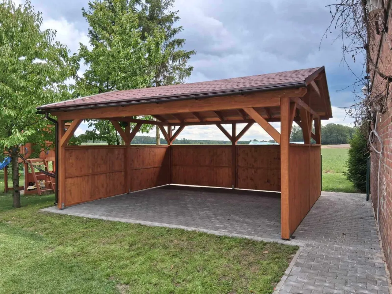 Drewniana wiata z brązowym dachem, idealna jako co na dach altany. Podjazd z kostki brukowej, otoczony zielenią.