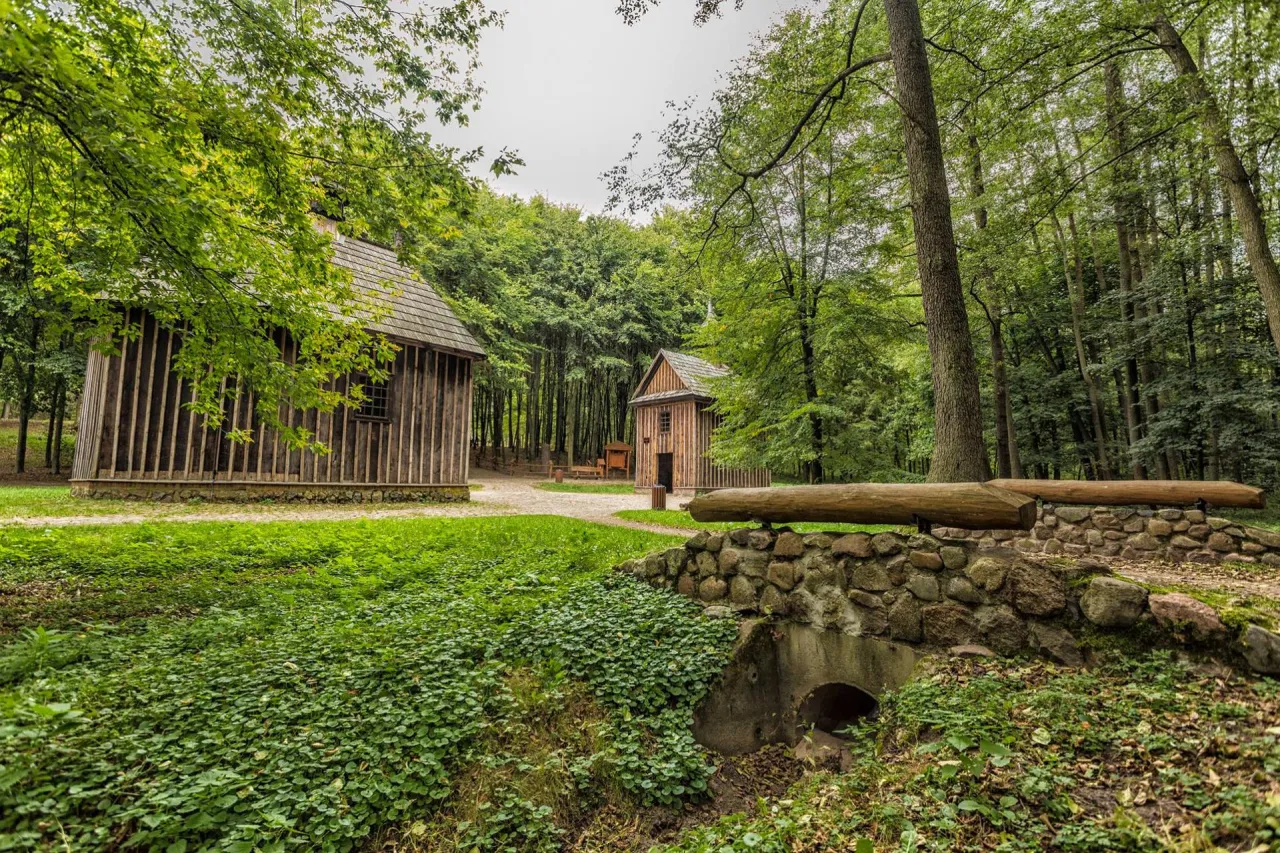 Drewniane chaty w rezerwacie przyrody Las Łagiewnicki, otoczone zielenią i lasem. Kamienny mostek z drewnianą balustradą.