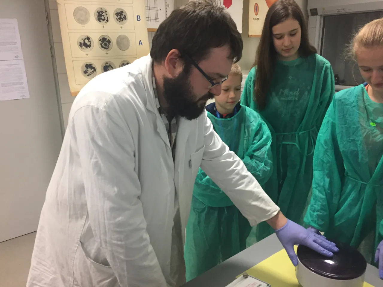 Nauczyciel w białym fartuchu pokazuje uczniom w zielonych fartuchach, czy praca w laboratorium jest trudna.