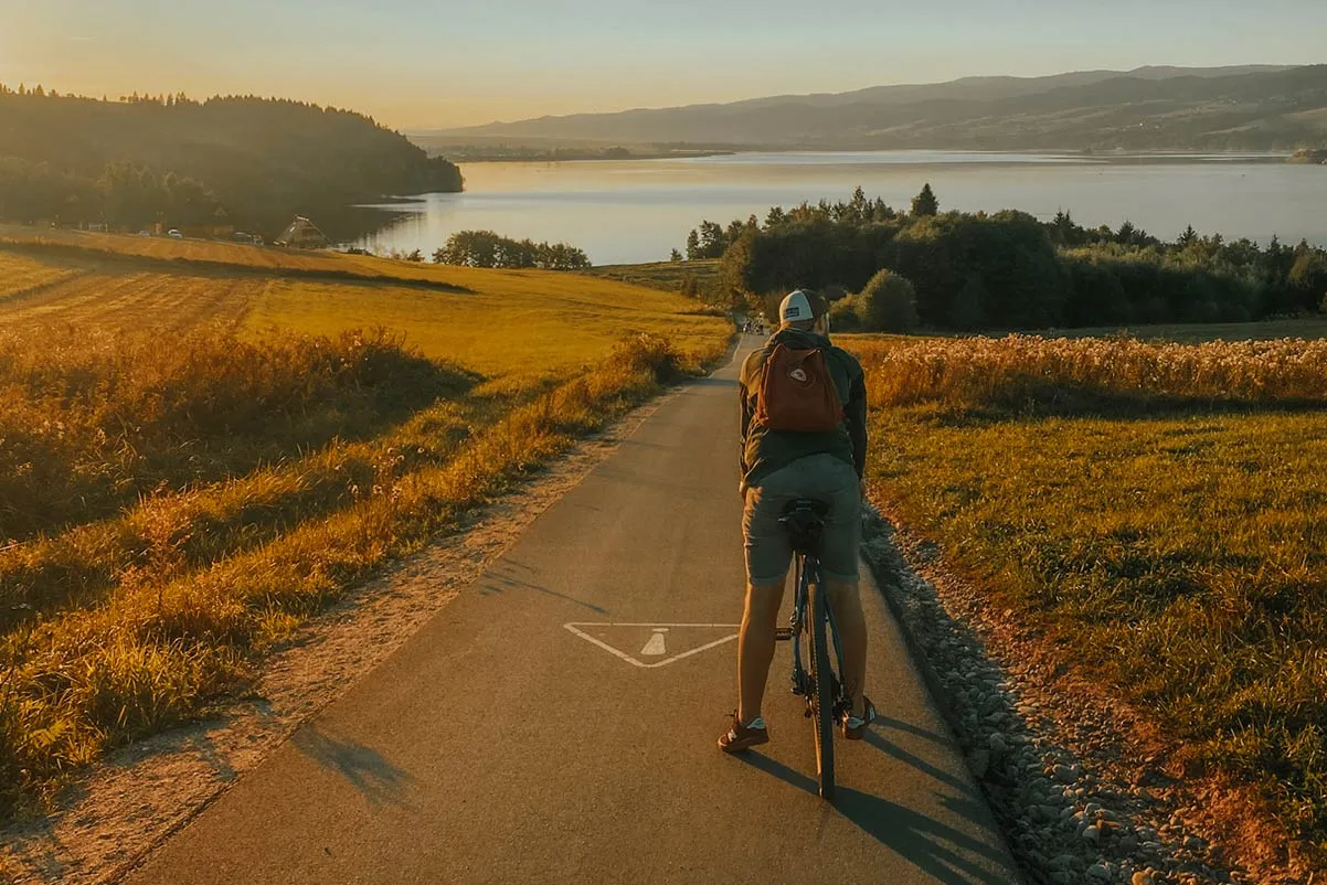 Rowerzysta na ścieżce rowerowej w Czorsztynie, złote światło zachodzącego słońca oświetla malowniczy krajobraz z jeziorem i wzg&oacute;rzami.