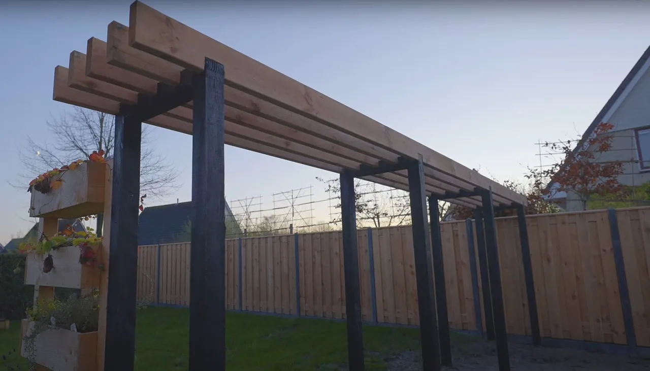 Jak zrobic pergole na dzialce? Zobacz, jak powstaje drewniana konstrukcja z czarnymi słupami i skrzynkami na rośliny.