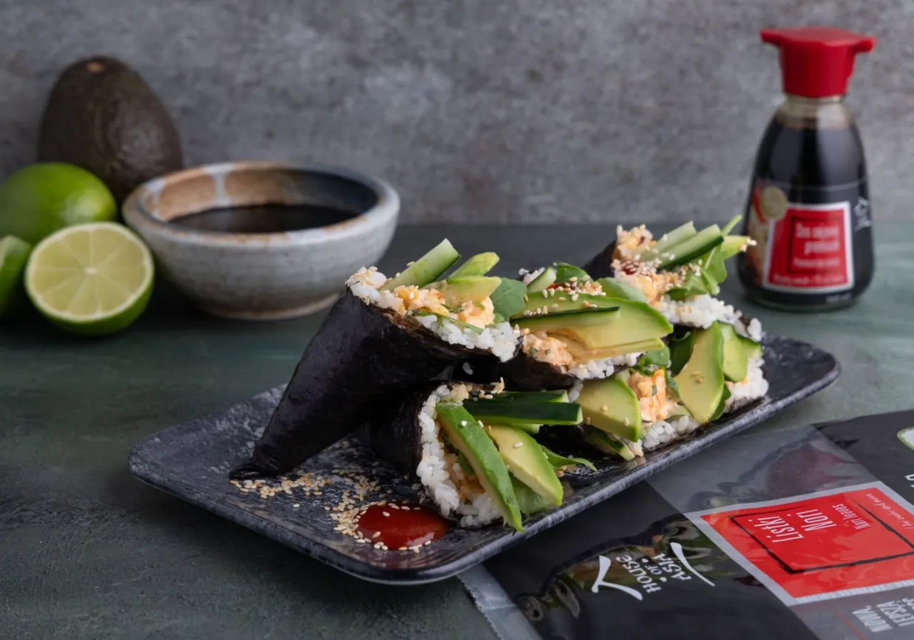 Szybkie sushi w formie rożk&oacute;w z awokado i og&oacute;rkiem, posypane sezamem. Obok sos sojowy, limonki i miseczka z sosem.