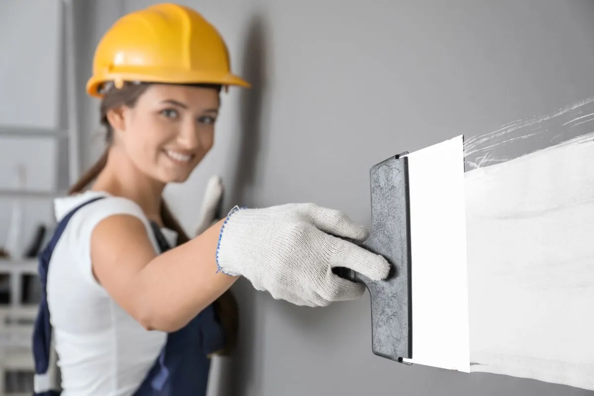 Uśmiechnięta budowlaniec w żółtym kasku nakłada gips na ścianę, sprawdzając, który gips szybciej schnie.