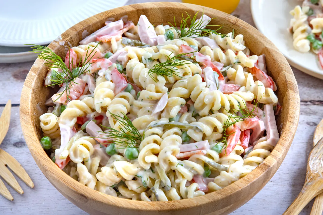 klassischer Nudelsalat in einer Schüssel auf einem Gartentisch mit Grillgut im Hintergrund