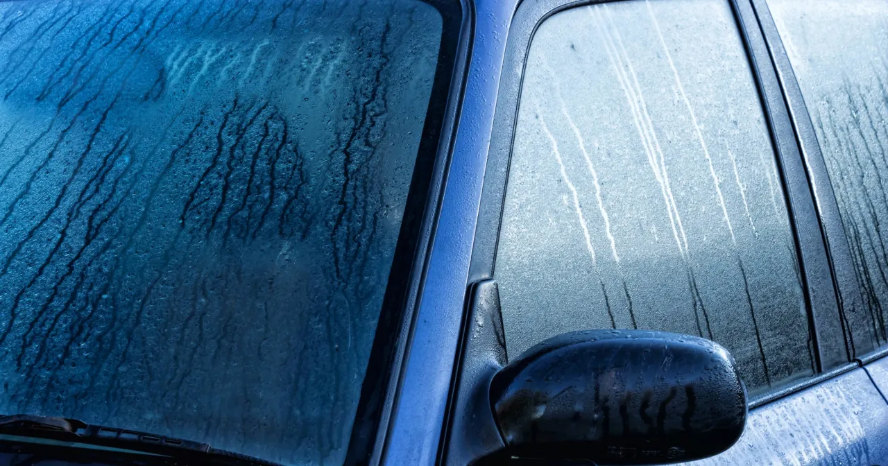 Krople wody spływają po zaparowanych szybach samochodu, tworząc smugi. Widoczny fragment lusterka bocznego.