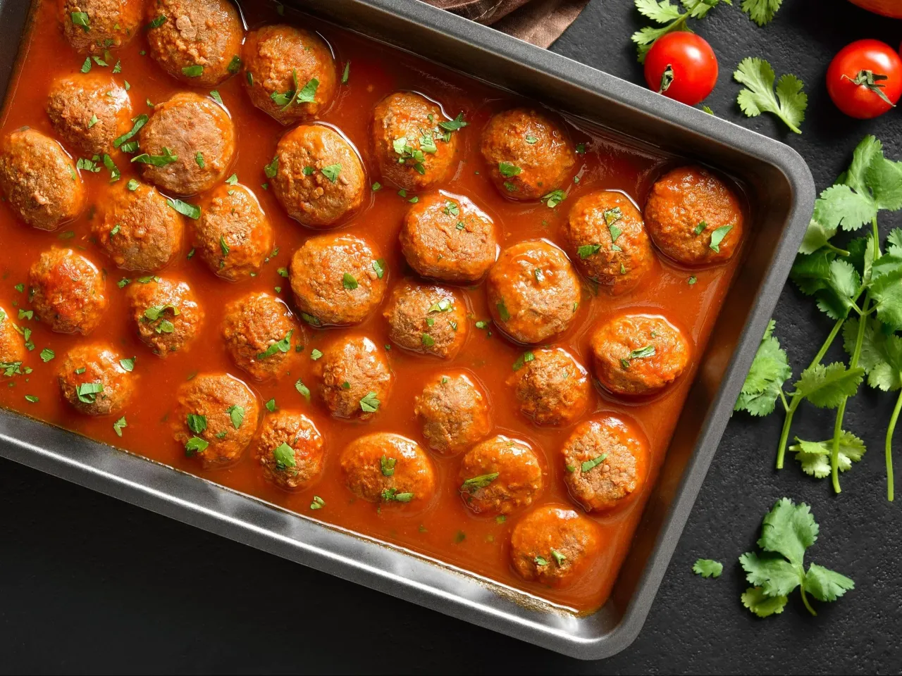 Pulpety w sosie pomidorowym, posypane natką pietruszki, w metalowej formie. Obok świeże pomidory i gałązki kolendry.