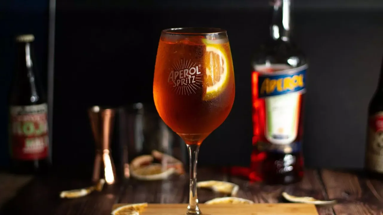 Orzeźwiający Aperol Spritz z plasterkiem pomarańczy, obok butelka Aperol. Idealny wybór zamiast Hugo.